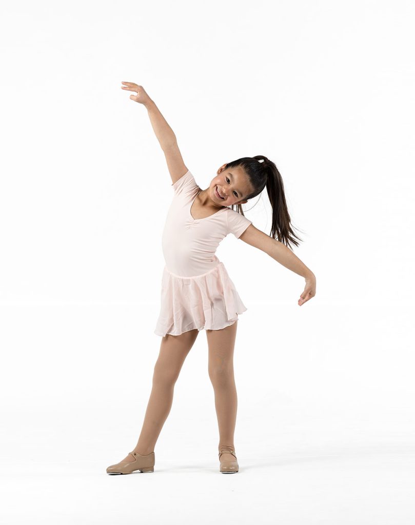 FullYear A young girl in a light pink dance dress and tan tap shoes poses with a joyful smile, leaning to the side with arms extended.